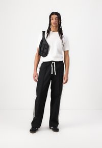 Black track pants with white side stripes, elastic waistband with drawstring; paired with a plain white t-shirt and a black shoulder bag.