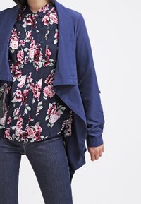 Giacca leggera blu navy con orlo asimmetrico, indossata sopra una blouse floreale blu scuro con rose rosa, abbinata a jeans scuri.