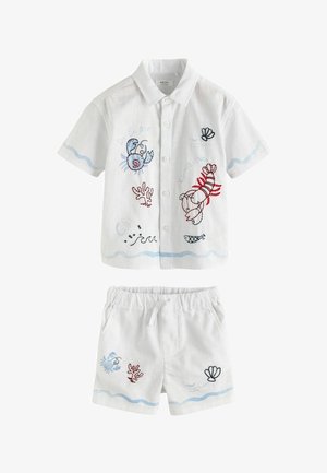 Ragazzo giovane in piedi, indossa una maglietta bianca e pantaloncini con ricami a tema marino in blu e rosso, e sandali blu.