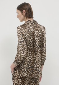 Donna di profilo che indossa una camicia a maniche lunghe con stampa leopardata e pantaloni coordinati, con le mani nelle tasche su uno sfondo neutro.
