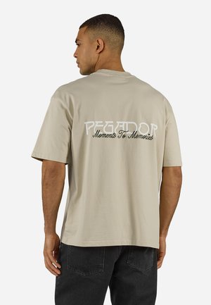 Άνδρας με κοντά μαλλιά φοράει μια μπεζ μπλούζα με το κείμενο «PEGADOR Moments To Memories» στην πλάτη, συνδυασμένη με μαύρο τζιν.