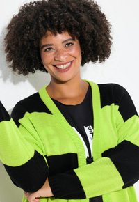 Cardigan a strisce verdi e nere con una trama a maglia larga e maniche lunghe. Caratterizzato da un collo a V e design aperto davanti.