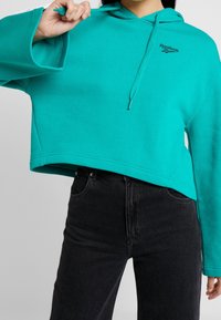Haut de survêtement turquoise court avec une capuche à cordon et un logo Reebok noir. Présente des manches larges et un tissu en mélange de coton doux. Associé à un jean noir.