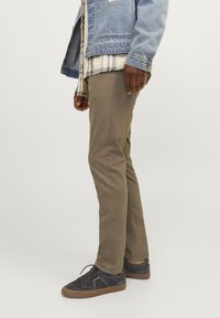 Uomo che indossa pantaloni slim-fit marroni, sneakers blu scure, una camicia a quadri e una giacca di denim blu, in piedi di lato su uno sfondo semplice.