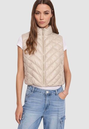 Donna che indossa un gilet beige trapuntato senza maniche sopra una camicia bianca a maniche corte e jeans in denim azzurro chiaro con la mano in tasca.