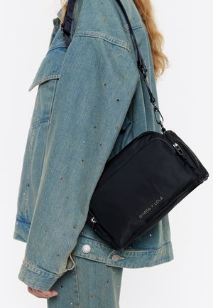 Schwarze Nylon-Crossbody-Tasche mit silbernem Reißverschluss und Branding, getragen über einer Jeansjacke mit verstreuten metallischen Verzierungen.