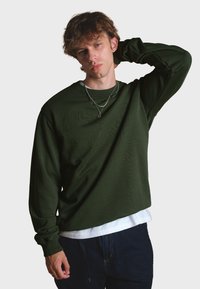 Sudadera de manga larga en verde oliva hecha de un tejido suave, con cuello redondo y un sutil detalle del logo, llevada sobre una camisa blanca.