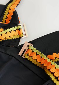 Čierny plavky z tkaniny s multikolorovým vlnitým lemom v oranžovej a zelenej farbe, s detailom z ružovo-zlatého kovu na zapínaní. Hladká textúra.