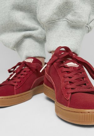 Zapatillas de ante rojo con una suela de goma texturizada marrón, con cordones a juego y detalles del logo. Vista en primer plano desde arriba.