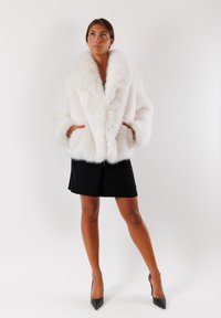 Cappotto in pelliccia sintetica bianca con un ampio colletto, chiusura doppiopetto e tasche laterali, abbinato a una gonna nera e scarpe con tacco alto.