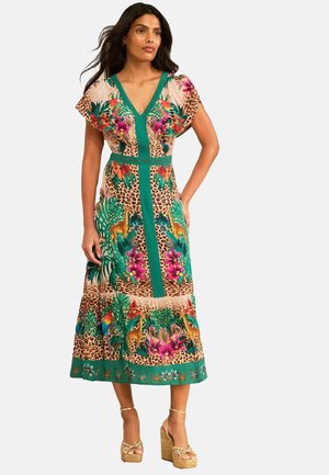 Frau trägt ein buntes Midi-Kleid mit V-Ausschnitt mit tropischen Blumen- und Tiermotiven, kombiniert mit beigen Plateau-Keilabsatzsandalen.