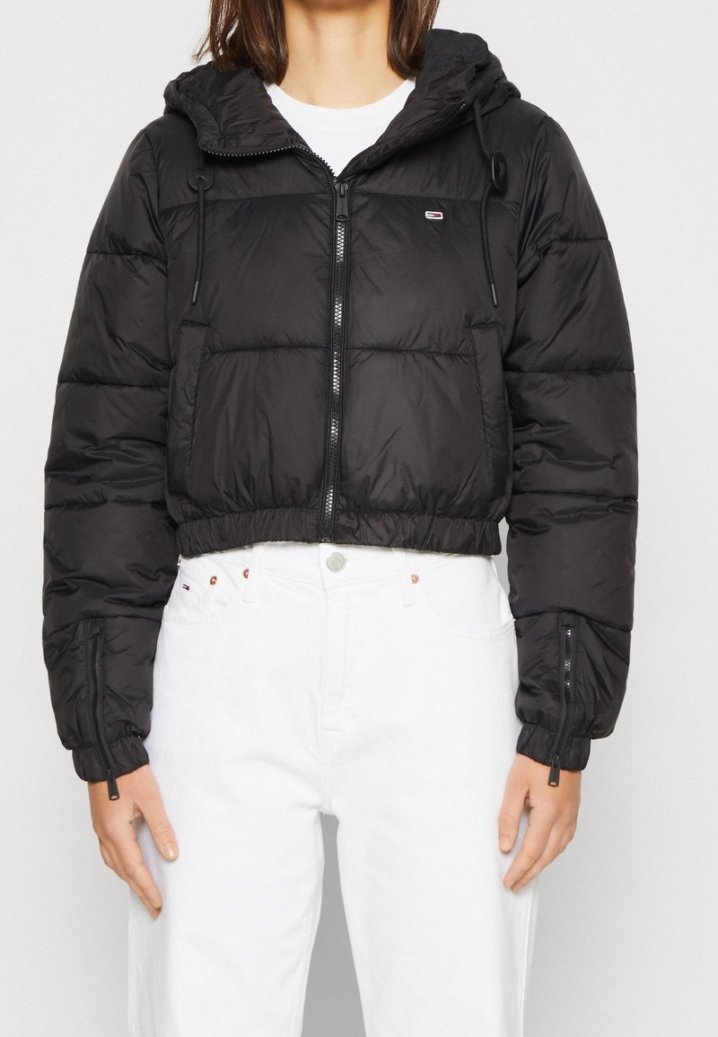 Veste courte matelassée noire à capuche, fermeture éclair frontale, poignets élastiques et détails à cordon de serrage. Le matériau semble brillant avec des sections matelassées.