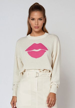 Frau trägt einen cremefarbenen Pullover mit einer großen pinken Lippen-Grafik, in einen hoch taillierten cremefarbenen Rock mit Gürtel und Taschen gesteckt.