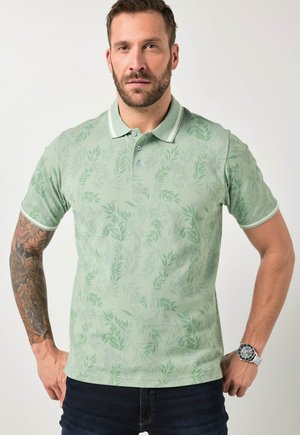 Mężczyzna w jasnooliwkowej koszulce polo z subtelnym liściastym nadrukiem, ciemnych dżinsach, z tatuażem na lewej ręce i srebrnym zegarkiem na prawej ręce na jednolitym tle.