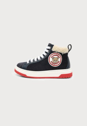 Czarny wysokowy sneaker z białymi sznurowadłami, czerwoną podeszwą, puszystym beżowym kołnierzem i okrągłą naszywką z niedźwiedziem i czerwonym napisem.