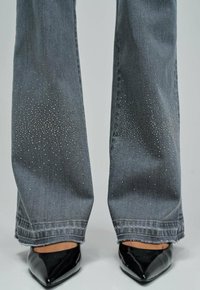 Grå jeans med små rhinestones utspridda vid fållen, parat med svarta lackade klackskor med spetsig tå, mot en ljus bakgrund.