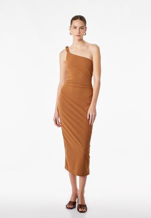 Frau trägt ein figurbetontes, einseitiges braunes Midikleid mit Knotendetail, goldene Creolen und dunkle offene High Heels vor weißem Hintergrund.