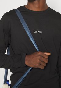 Schwarzes Langarmshirt mit dem weißen "LES DEUX"-Logo. Die Person trägt eine blaue Umhängetasche über der Schulter.