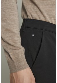 Svarta byxor med en slät yta och en liten lyster, med en sidoficka och en liten logotypdetalj. Klädseln kompletteras av en beige stickad tröja.