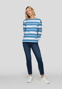 Blau-weißer gestreifter Langarm-Top mit Textakzenten, kombiniert mit dunklen Jeans und weißen Sneakern. Glatter Stoff, lockere Passform, Rundhalsausschnitt.