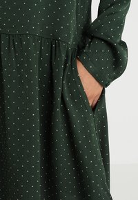 Vestido verde oscuro con lunares blancos. Hecho de una tela suave. Presenta mangas largas y una cintura fruncida. Incluye un bolsillo lateral.