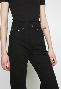 Calças de ganga pretas com uma cintura alta, apresentando cinco bolsos, metais prateados e um design ajustado. O tecido tem uma textura suave e mate.