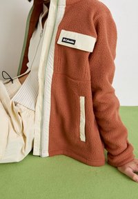 Brązowa polarowa kurtka z kremowymi akcentami, z przodu na zamek błyskawiczny i kieszonką na piersi. Posiada teksturowany materiał oraz luźny krój.