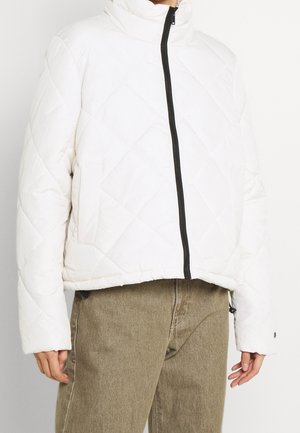 Personne portant une veste matelassée blanche avec une fermeture éclair noire et un pantalon marron clair, visible des épaules aux hanches sur un fond uni.