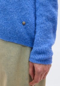 Κοντινό πλάνο ενός χεριού που ακουμπά στο πλάι, φοράει ένα χνουδωτό μπλε πλεκτό πουλόβερ με ένα μικρό μεταλλικό σήμα λογότυπου και μπεζ παντελόνι.