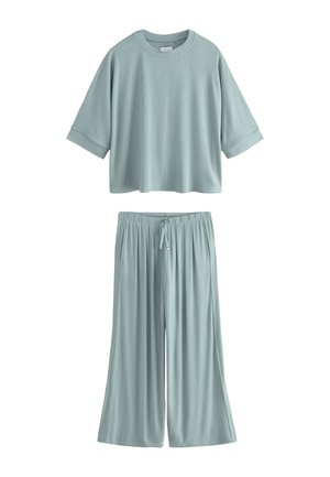 Lichtblauwe geribde stoffen top en broek met een klein metalen merkteken op het topje dicht bij de zoom, met een zichtbare linkerarm en hand.