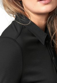 Schwarze Kragenbluse aus glattem Stoff, mit einer Knopfleiste und einer sauberen Verarbeitung, die sichtbare Nähte entlang der Säume aufweist.