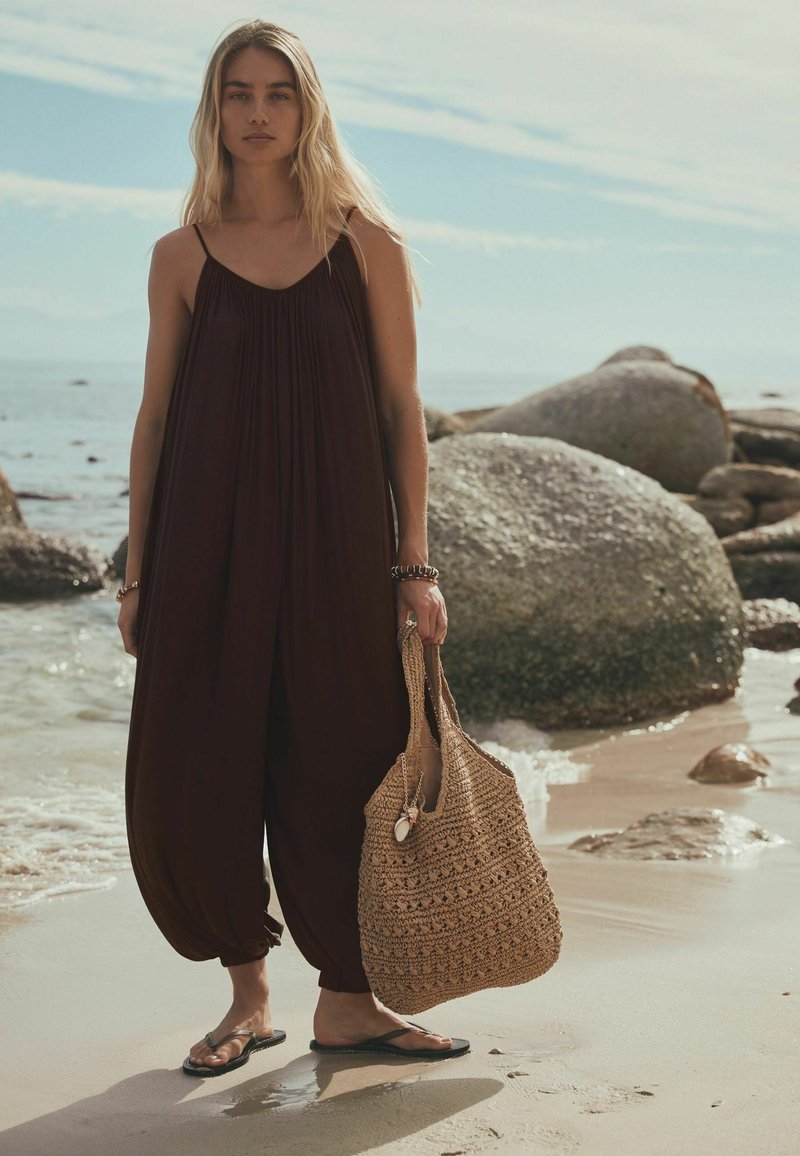 Jeune femme en combinaison ample noire tenant un sac de plage tressé, debout pieds nus sur une plage de sable avec des rochers et la mer calme en arrière-plan.