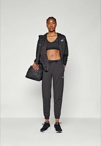 Black athletic outfit with a zip-up hoodie, sports bra, and tapered joggers. Black sneakers and a black sports bag complete the look.