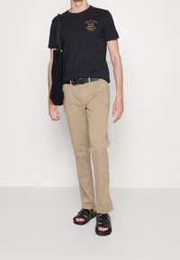 Schwarzes Baumwoll-T-Shirt mit gelbem Text, beige Hosen und schwarzen Sandalen. Das Outfit umfasst eine schwarze Tasche, die über eine Schulter geschlungen ist.