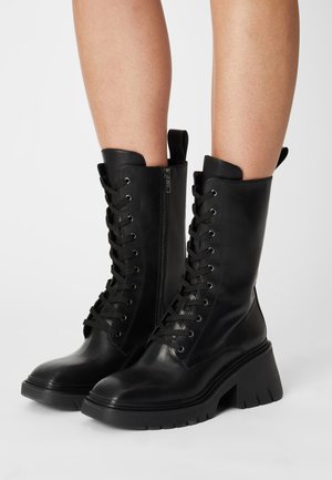 Bottes de combat en cuir noir mi-mollet avec semelles crantées épaisses, lacets à l'avant, fermeture éclair latérale et boucles de traction, portées sur des jambes nues contre un fond clair.
