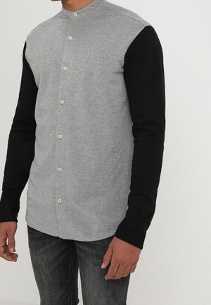 Mann trägt ein graues Hemd mit Knöpfen und schwarzen langen Ärmeln sowie dunkle Jeans, steht vor einem schlichten hellen Hintergrund.