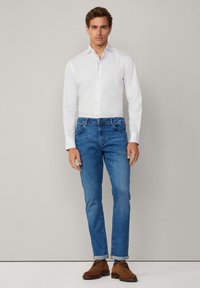 Modèle masculin portant une chemise blanche à boutons et un jean en denim bleu. Le jean a une coupe droite, avec des ourlets retroussés et des chaussures habillées marron.