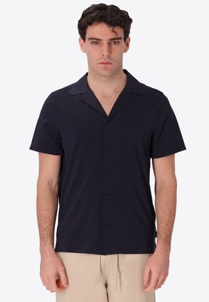 Uomo che indossa una camicia nera a maniche corte con bottoni e pantaloni beige, in piedi davanti a uno sfondo chiaro e semplice.