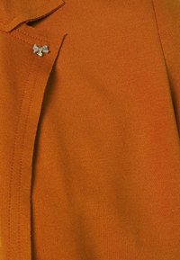 Neulepusero oranssia, jossa on kaulus ja koristeellinen röyhelökoristus kristallilla. Sujuva rakenne näkyvin ompelutaisin.