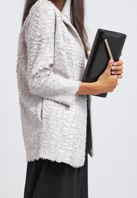 Teksturoitu vaalea harmaa takki, jossa on väljä istuvuus ja kerroksellinen kuvio. Kädessä pidetään tyylikästä mustaa clutch-laukkua, jossa on metallinen yksityiskohta.