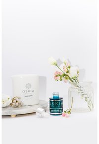 Vit ljus med etiketten "OSKIA ROSE DE MAI", blå glasflaska med pipett, marmorbasis, klar glasvas, mjuka rosa och vita blommor.