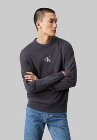 Σκούρο γκρι φούτερ με ραμμένο λαιμό και μανίκια. Διαθέτει λευκό λογότυπο "CK" και άνετη εφαρμογή. Φοριέται πάνω από ένα μπλε t-shirt και τζιν παντελόνι.