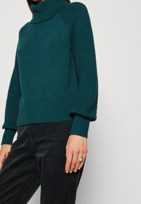 Teal knitted sweater with a high collar. Features a textured pattern on the sleeves and ribbed cuffs and hem. Paired with dark corduroy pants.