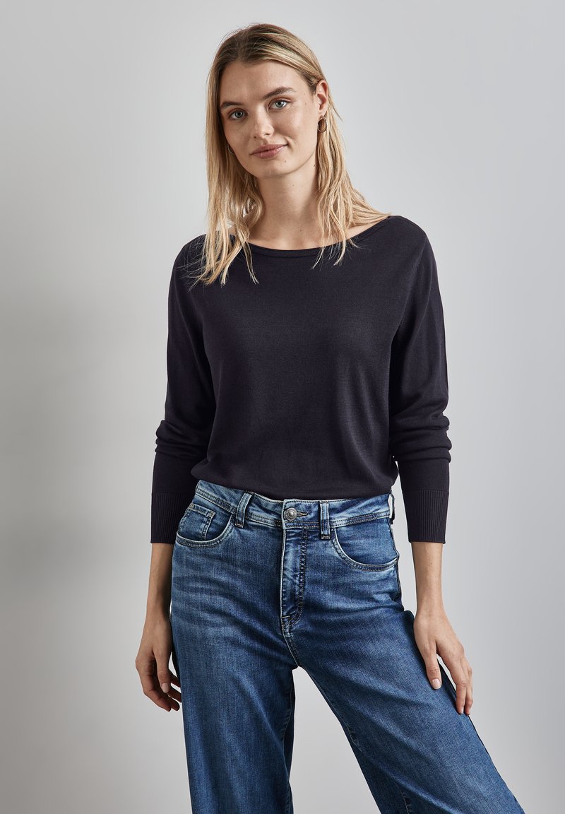 Haut noir en tricot à manches longues avec un col large, associé à un jean bleu à jambe large. Les deux vêtements présentent une texture lisse et un design décontracté.
