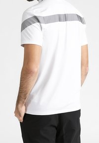 Bílá polo košile s krátkým rukávem s šedým pruhovaným vzorem přes ramena. Vyrobena z hladkého a lehkého materiálu.