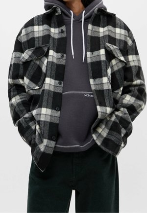 Person trägt eine schwarz-weiß karierte, zum Knöpfen vorgesehene Jacke über einem dunkelgrauen Hoodie mit weißen Kordeln, die Hände in den Jackentaschen.