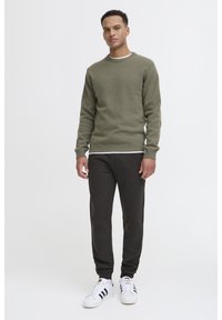 Olijfgroen trui met ronde hals, ribgebreide manchetten en zoom, gecombineerd met donkere sweatpants en witte sneakers met zwarte strepen.