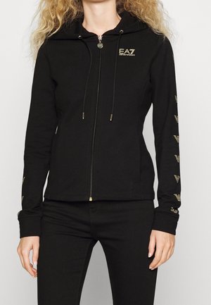Kobieta nosząca czarną bluzę z kapturem na suwak z złotymi logo EA7 Emporio Armani na piersi i rękawach, w połączeniu z czarnymi spodniami.
