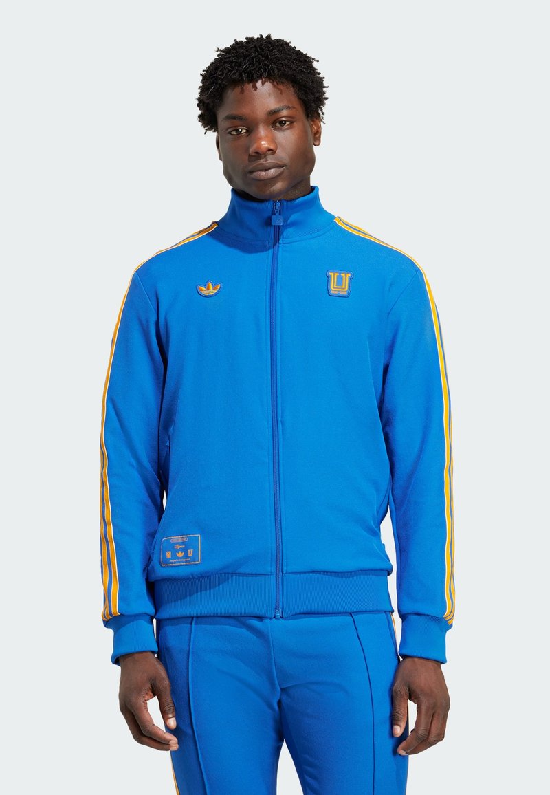 Blå zip-up jakke med gule sideløbende striber, forlommer og broderede logoer. Blødt stof med en tætsiddende pasform og ribkantede manchetter.