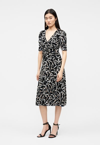 Γυναίκα που στέκεται φορώντας μαύρο midi φόρεμα με λευκό και μπεζ σχέδιο φύλλων, με σούρες μπροστά, φουσκωτά μανίκια και μαύρα ψηλοτάκουνα με λουράκι στον αστράγαλο, σε λευκό φόντο.